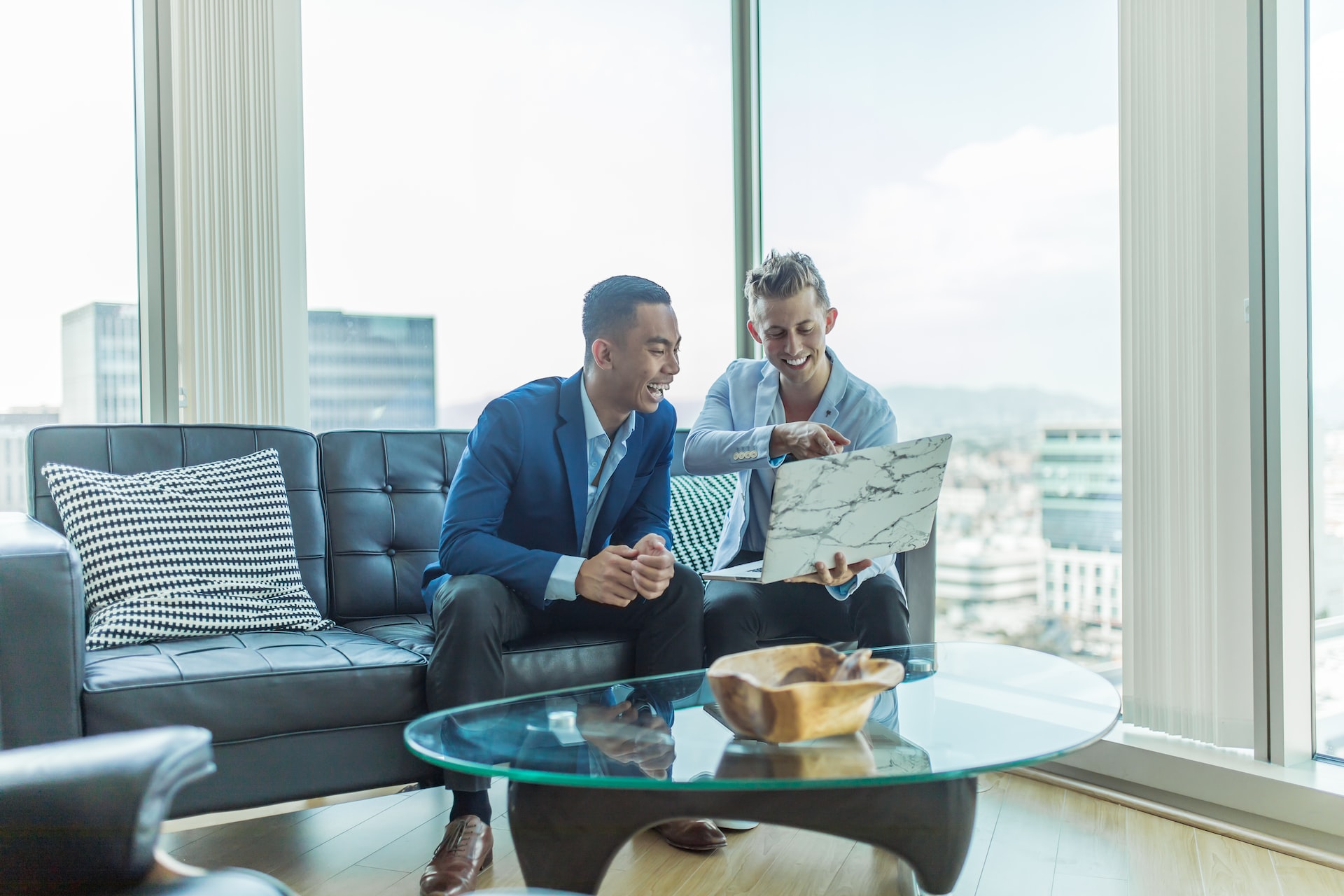 two men in suits sitting on couch smiling at laptop two men in suits sitting on couch smiling at laptop
