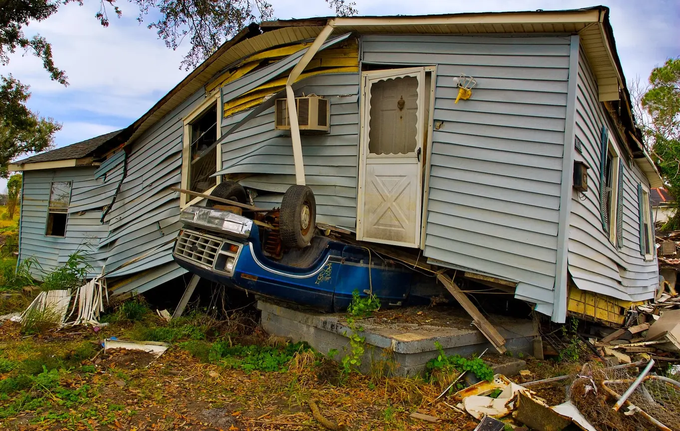 A house struck by a disaster and upended, with a car under it