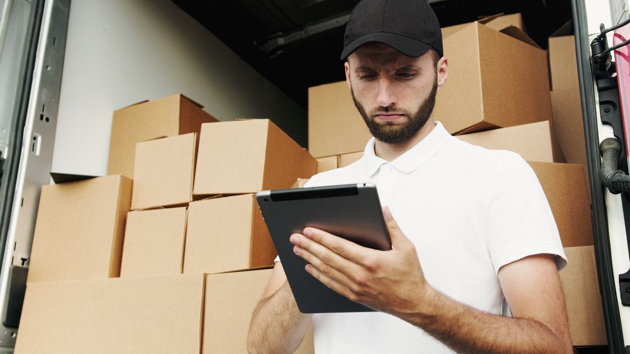 Lieferant mit Mütze schaut auf Tablet mit Kisten im Hintergrund Lieferant mit Mütze schaut auf Tablet mit Kisten im Hintergrund