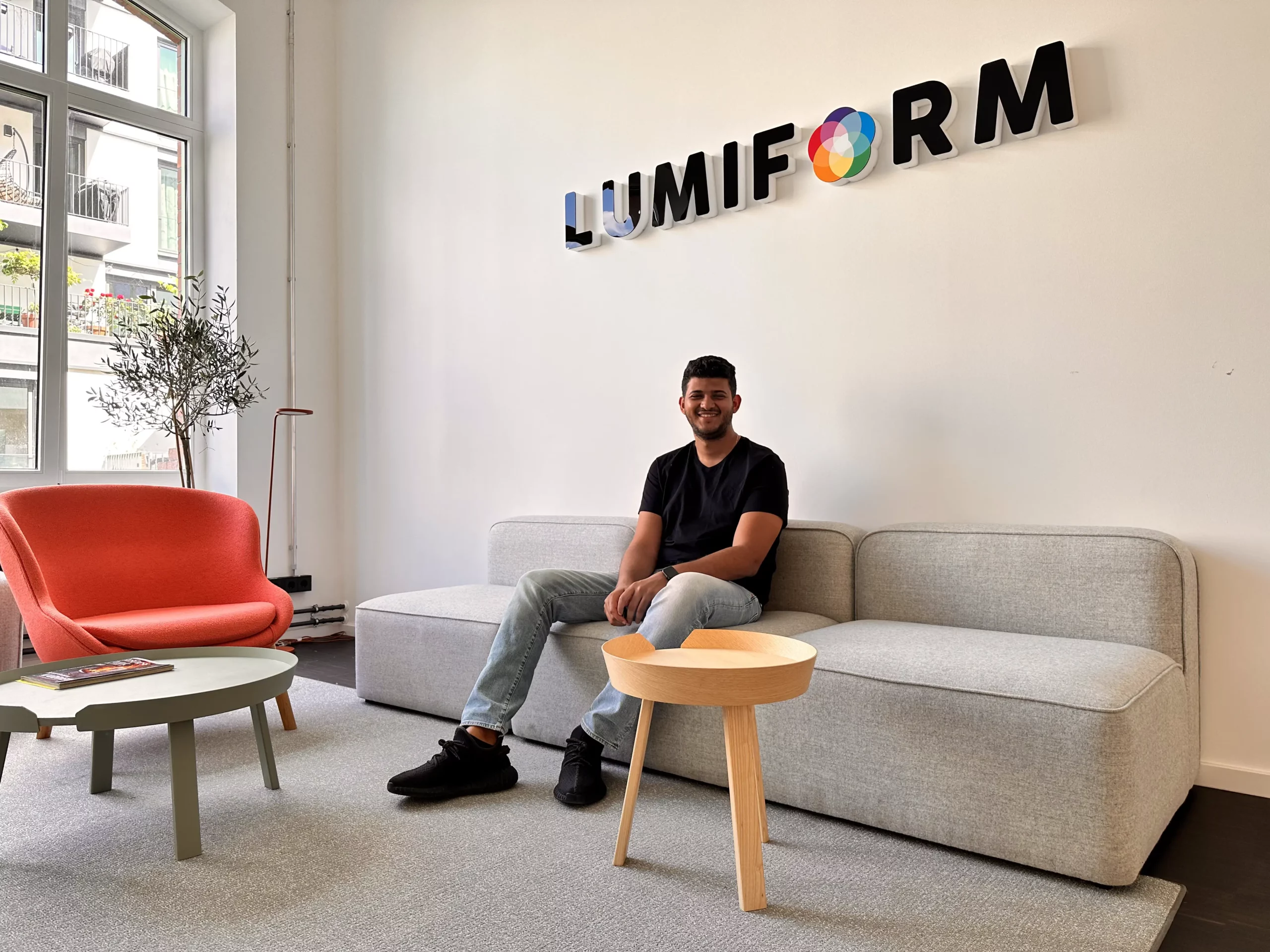 Ein junger Mann sitzt auf einer grauen Couch, hinter ihm ein Logo auf dem Lumiform steht